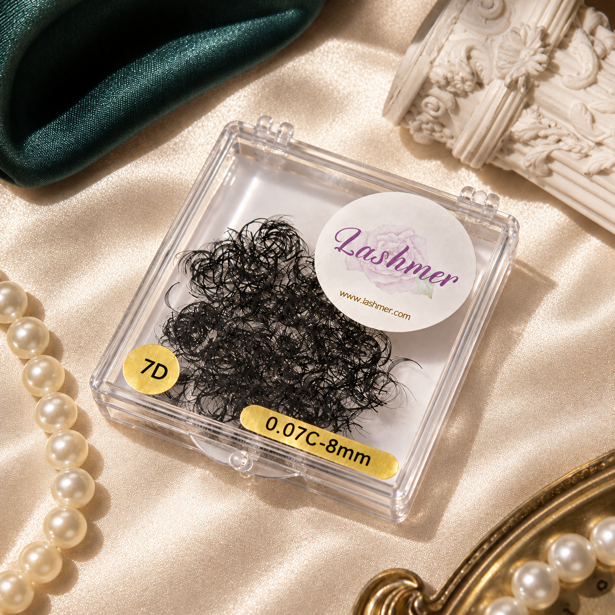 A clear square plastic case containing black eyelash clusters, placed on a champagne satin surface with decorative pearls, a green velvet fabric, and ornate white frame pieces around it. The case has a round white sticker reading 'Lashmer' and a yellow sticker reading '7D'.